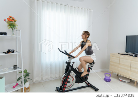 Young happy woman pedals spin bike for home fitness activity, people enjoying healthy exercise inside bright room displaying white curtains and shelf with kettlebell 132374839