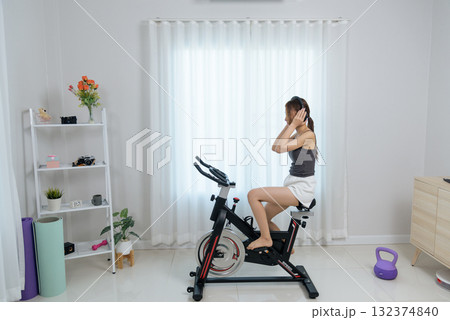 Young woman conducts home fitness activity, exercising indoors on stationary bike, wearing headphones for invigorating workout, surrounded by various health equipment 132374840