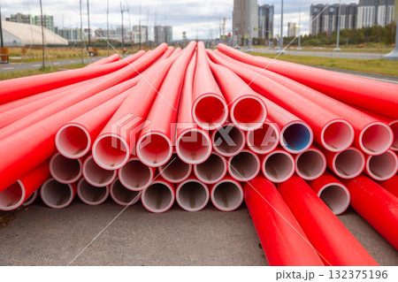 Stack of bright red plastic conduit pipes is arranged in a neat stack on a construction site. Modern buildings rise in the background under a partly cloudy sky, indicating urban development. Stack of bright red plastic conduit pipes is arranged in a neat stack on a construction site. Modern buildings rise in the background under a partly cloudy sky, indicating urban development. 132375196