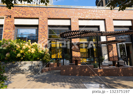 A cozy outdoor seating area of cafe or restaurant features stylish furniture under a canopy in a vibrant urban environment. Lush plants and flowers enhance the inviting atmosphere on a sunny day. 132375197