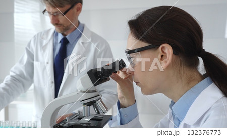 Female scientist wearing protective glasses looking through a microscope during medical research in laboratory with her male colleague working in the background. Medicine and science concept 132376773
