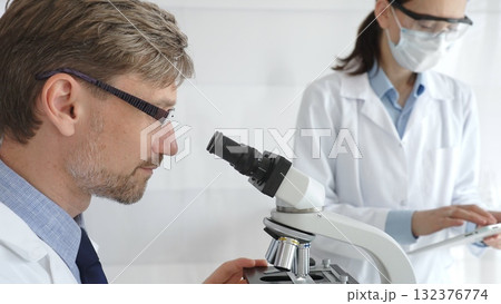 A male researcher examines a sample under a microscope, a female lab technician in a protective mask stands nearby, both in white lab coats during the study. Medicine and Science 132376774