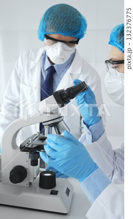Two scientists in lab coats, face masks, and protective glasses conducting research with a microscope in a laboratory. Medicine and science concept 132376775