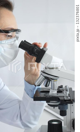 A female scientist in a protective mask and glasses carefully examines a sample under a microscope while conducting research in a modern laboratory. Concept of medicine and science A female scientist in a protective mask and glasses carefully examines a sample under a microscope while conducting research in a modern laboratory. Concept of medicine and science 132376801