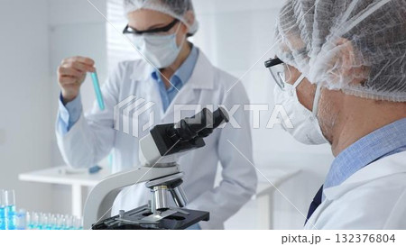A male scientist wearing a protective mask, glasses and hairnet works with a microscope while a female colleague helps him by holding a tablet and taking notes in a sterile laboratory. Concept of A male scientist wearing a protective mask, glasses and hairnet works with a microscope while a female colleague helps him by holding a tablet and taking notes in a sterile laboratory. Concept of 132376804