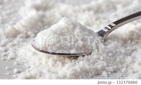 A close-up of a spoon holding a heap of coarse white sea salt crystals, with a powdery texture evident. 132378860
