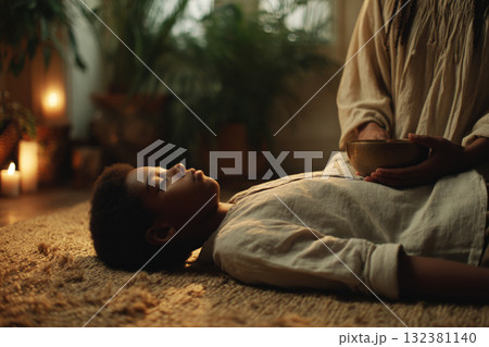 A child lies on a soft surface, eyes closed, as an adult gently places their hands on the child's head, surrounded by lit candles and plants, creating a peaceful atmosphere A child lies on a soft surface, eyes closed, as an adult gently places their hands on the child's head, surrounded by lit candles and plants, creating a peaceful atmosphere 132381140