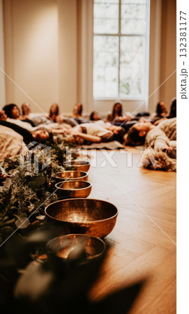 In a tranquil room filled with greenery, participants lie on mats while a facilitator leads a sound therapy session using singing bowls and candles for ambiance In a tranquil room filled with greenery, participants lie on mats while a facilitator leads a sound therapy session using singing bowls and candles for ambiance 132381177