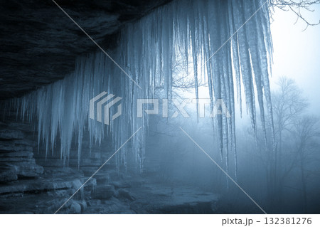 Icicles of various sizes dangle from the rocky cliffs in a forest during winter. The ground is covered in ice and there is a serene blue hue among the frozen landscape 132381276