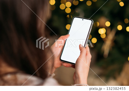 Close-up of woman using smartphone with blank white screen near Christmas tree with golden festive lights. 132382455
