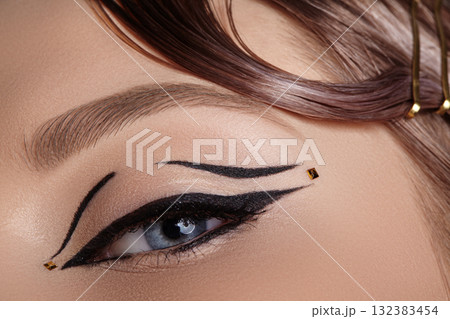 Close-up macro of a closed female eye with black winged eyeliner and tiny gold dot embellishment, soft lighting highlighting lash curve and eyelid decoration for editorial beauty use 132383454