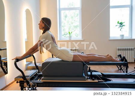 Woman doing Swan exercise on Pilates Reformer for the chest and back muscles, focusing on flexibility, strength, and control with determination Woman doing Swan exercise on Pilates Reformer for the chest and back muscles, focusing on flexibility, strength, and control with determination 132384915