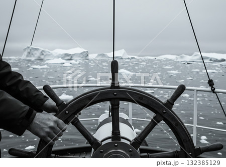 Hands on a ship's wheel steering through an icy sea full of icebergs. Polar exploration and navigation concept. Monochrome photo of an arctic adventure 132385129