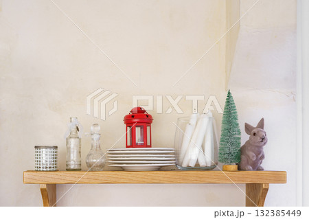 Wooden shelf with candles, red lantern, glass bottles, plates, and decorative Christmas bunny Wooden shelf with candles, red lantern, glass bottles, plates, and decorative Christmas bunny 132385449