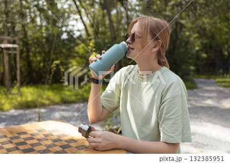 Young woman drinking water from reusable bottle while sitting outdoors at wooden table Young woman drinking water from reusable bottle while sitting outdoors at wooden table 132385951