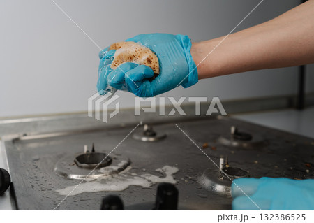 Housekeeper wearing blue gloves squeezing soapy water from sponge during cleaning dirty gas stove, to removing stubborn grime and grease. Concept of domestic work, housekeeping and household. Housekeeper wearing blue gloves squeezing soapy water from sponge during cleaning dirty gas stove, to removing stubborn grime and grease. Concept of domestic work, housekeeping and household. 132386525