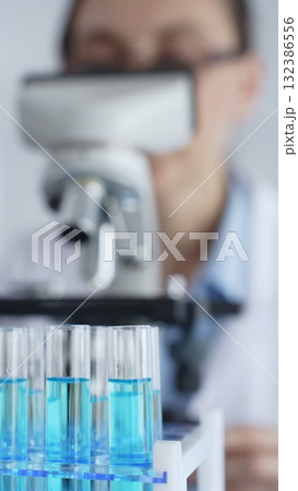 Close up of test tubes with blue liquid while scientist focusing on analyzing samples under a microscope in a laboratory setting. Medicine, healthcare and science concept 132386556