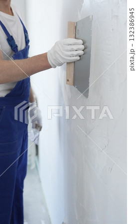 Man construction worker wearing protective gloves and blue construction coveralls, spreading plaster smoothly across wall using professional drywall taping knife 132386945