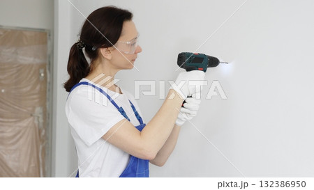 Professional female worker wearing blue overall, safety glasses and gloves, is using cordless drill to make hole in white wall during renovation work. Portrait view 132386950