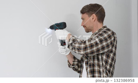 Male construction worker in checked shirt and safety gloves is screwing a screw into the wall using a cordless screwdriver, carrying out a careful home renovation. Portrait view Male construction worker in checked shirt and safety gloves is screwing a screw into the wall using a cordless screwdriver, carrying out a careful home renovation. Portrait view 132386958