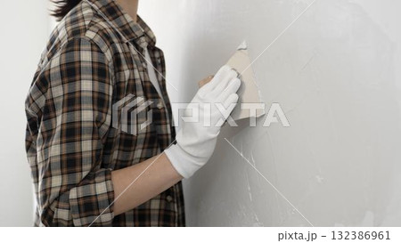 Unknown female construction worker wearing protective gloves and checkered shirt is spreading plaster on white wall during renovation project, smoothing surface with putty knife 132386961