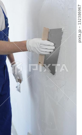 Man construction worker wearing protective gloves and blue construction coveralls, spreading plaster smoothly across wall using professional drywall taping knife 132386967