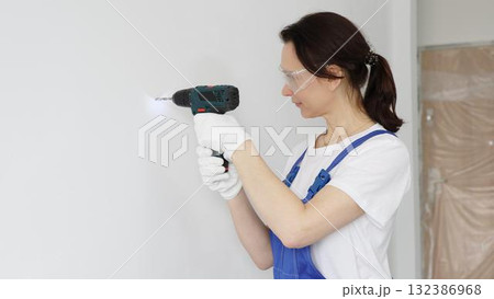 Professional female worker wearing blue overall, safety glasses and gloves, is using cordless drill to make hole in white wall during renovation work. Portrait view 132386968