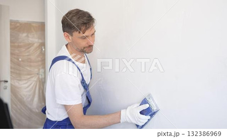 Man construction worker wearing protective white gloves and blue construction coveralls, is sanding white wall with blue hand block, preparing smooth surface during interior home renovation project 132386969