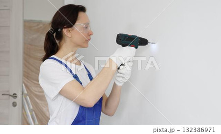 Woman worker wearing blue overall, safety glasses and gloves, is using cordless drill to make hole in white wall during renovation work. Horizontal portrait view 132386971