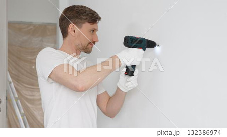 Professional male worker wearing white t-shirt and protective gloves, is drilling hole in a wall with cordless drill during home renovation work. Horizontal portrait view Professional male worker wearing white t-shirt and protective gloves, is drilling hole in a wall with cordless drill during home renovation work. Horizontal portrait view 132386974