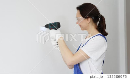 Professional female worker wearing blue overall, safety glasses and gloves, is using cordless drill to make hole in white wall during renovation work. Portrait view 132386976