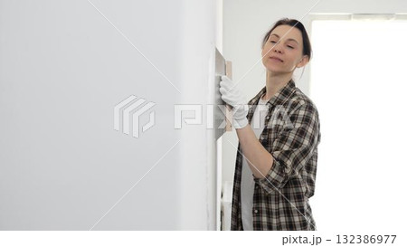 Female construction professional wearing checkered shirt is smoothing white wall surface, applying putty with professional spatula technique, showcasing skilled interior renovation work Female construction professional wearing checkered shirt is smoothing white wall surface, applying putty with professional spatula technique, showcasing skilled interior renovation work 132386977