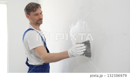 Middle aged male construction worker wearing gloves and blue overalls is using a putty knife to apply plaster on a white wall, performing home renovation 132386979
