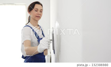 Professional female construction worker wearing blue coveralls is using a finishing trowel to carefully apply plaster on a wall, demonstrating expertise in home renovation 132386980