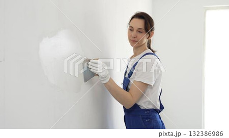 Construction worker woman spreading finishing putty on white wall, wearing protective gloves while smoothing surface with putty knife for precise home repair 132386986