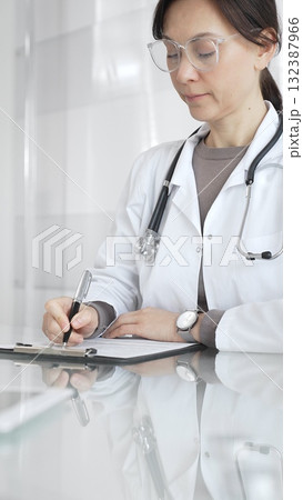 Female doctor in white coat and glasses writing medical notes on a clipboard at clinic desk, managing patient records. Medicine and health care Female doctor in white coat and glasses writing medical notes on a clipboard at clinic desk, managing patient records. Medicine and health care 132387966
