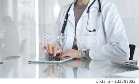 Physician in lab coat with stethoscope reviewing patient data on a tablet in a bright modern clinical office via touchscreen. Medicine and health care 132388082
