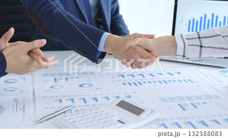Business people shaking hands over financial documents and laptop, confirming successful partnership, agreement, or merger in a corporate office meeting 132388083