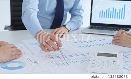 Business professional pointing at printed charts with a pen during a meeting, laptop showing bar graphs and calculator on table while team collaborates on financial analysis and planning 132388088