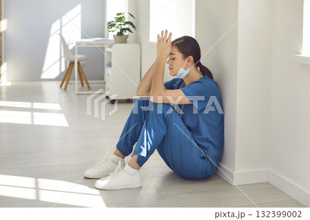 Tired stressed woman doctor wearing blue face mask and uniform sitting on hospital floor. 132399002
