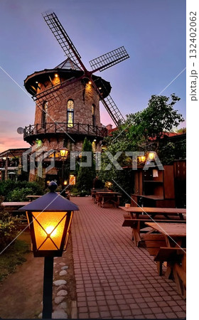 A stone building shaped like a windmill stands illuminated against a colorful twilight sky. It is not doing anything. The foreground shows an empty outdoor patio with wooden tables, benches, and warm, 132402062