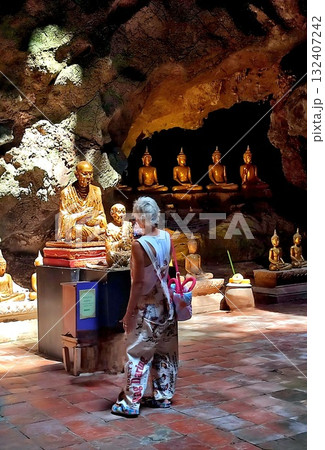 Tourist woman in patterned overalls stands inside a dimly lit Thai cave temple, looking at golden Buddha statues. Tourist woman in patterned overalls stands inside a dimly lit Thai cave temple, looking at golden Buddha statues. 132407242