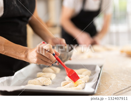 Female hands brushing uncooked dough pieces on baking sheet with oil before baking in oven closeup Female hands brushing uncooked dough pieces on baking sheet with oil before baking in oven closeup 132407911