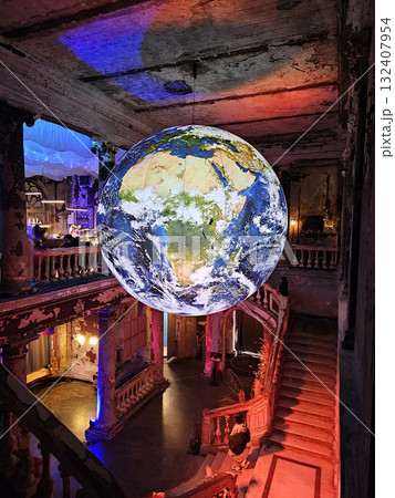 A large, illuminated globe of Earth hangs in a derelict historic hall with crumbling walls and old staircases, glowing brightly. 132407954