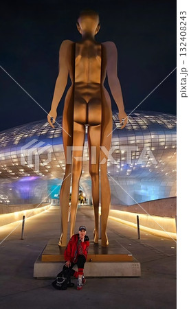 A stylish person sits under a massive golden human-like art sculpture outside the futuristic, curved Dongdaemun Design Plaza at night. A stylish person sits under a massive golden human-like art sculpture outside the futuristic, curved Dongdaemun Design Plaza at night. 132408243