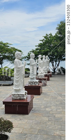 A row of white, praying Arhat statues on red pedestals stands proudly on a paved terrace, surrounded by lush green trees. A row of white, praying Arhat statues on red pedestals stands proudly on a paved terrace, surrounded by lush green trees. 132408300