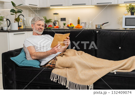 Middle-aged man enjoys reading book wrapped in plaid smiles and relaxes during quiet leisure time 132409074