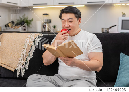 Asian man bites red apple enjoys juicy taste and read short health tips in book with calm smile well Asian man bites red apple enjoys juicy taste and read short health tips in book with calm smile well 132409098