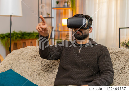 Indian man on sofa wears VR headset at home and reacts with excitement to immersive virtual world 132409587