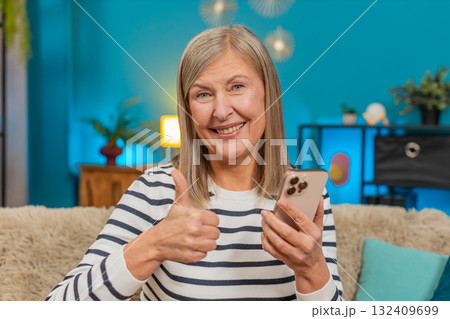 Mature woman viewing social media photos on smartphone, smiling, giving thumbs up approval at home Mature woman viewing social media photos on smartphone, smiling, giving thumbs up approval at home 132409699